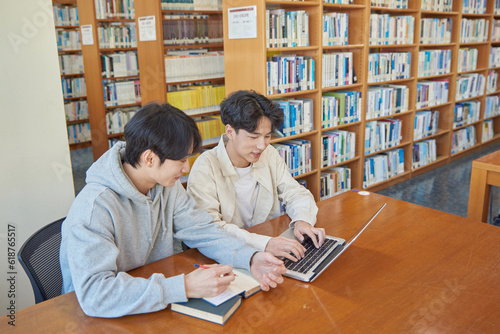 A male student sits at a desk in a university library in South Korea, Asia, looking at a laptop computer with bookshelves of books in the background.
