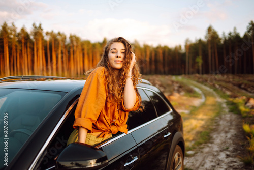 Beautiful traveler woman is resting and enjoying the sunset in the car. Happy woman on a summer trip, travels leaning out of the car window. Active lifestyle, tourism. Travel concept.