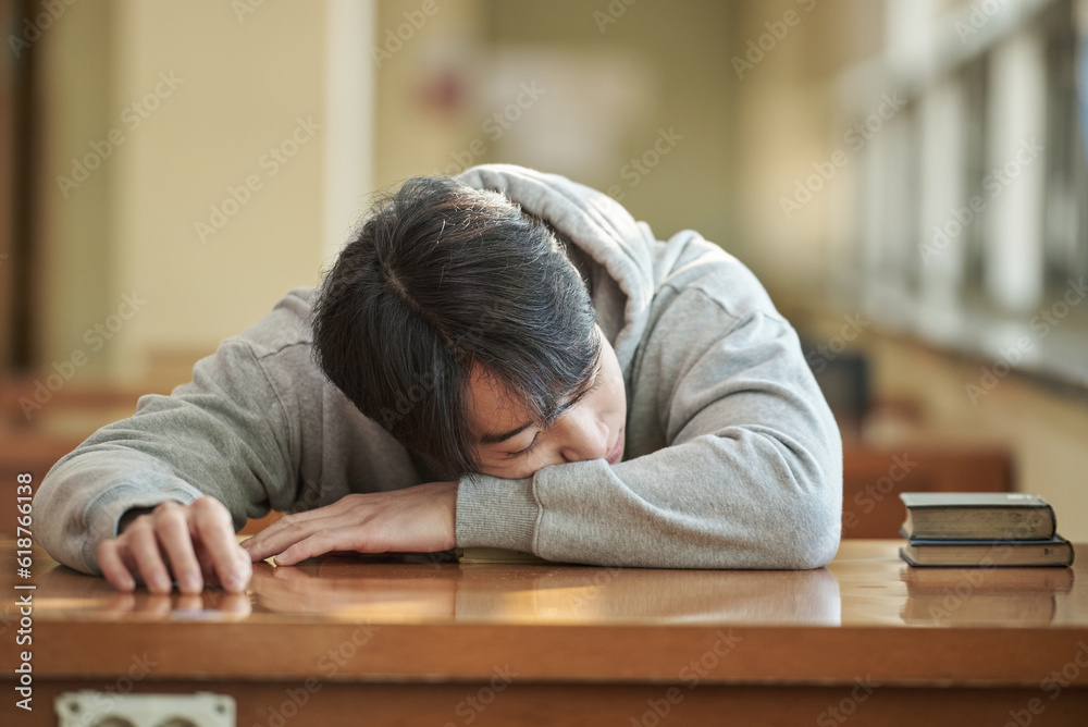 Fototapeta premium Asian Korean University Library Young male college student model sitting at a desk by a window with light coming in, reading a book, looking out the window or leaning down.