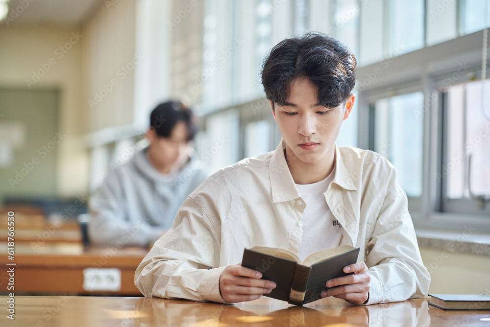 Three college students, both male and female, are sitting or standing at a desk in a library at a South Korean university in Asia, looking at laptops and books while having a conversation. Three college students, both male and female, are sitting or standing at a desk in a library at a South Korean university in Asia, looking at laptops and books while having a conversation.