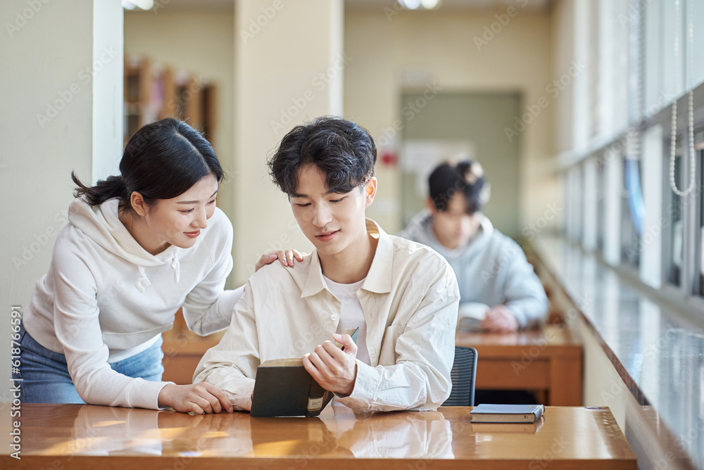 Three college students, both male and female, are sitting or standing at a desk in a library at a South Korean university in Asia, looking at laptops and books while having a conversation. Three college students, both male and female, are sitting or standing at a desk in a library at a South Korean university in Asia, looking at laptops and books while having a conversation.