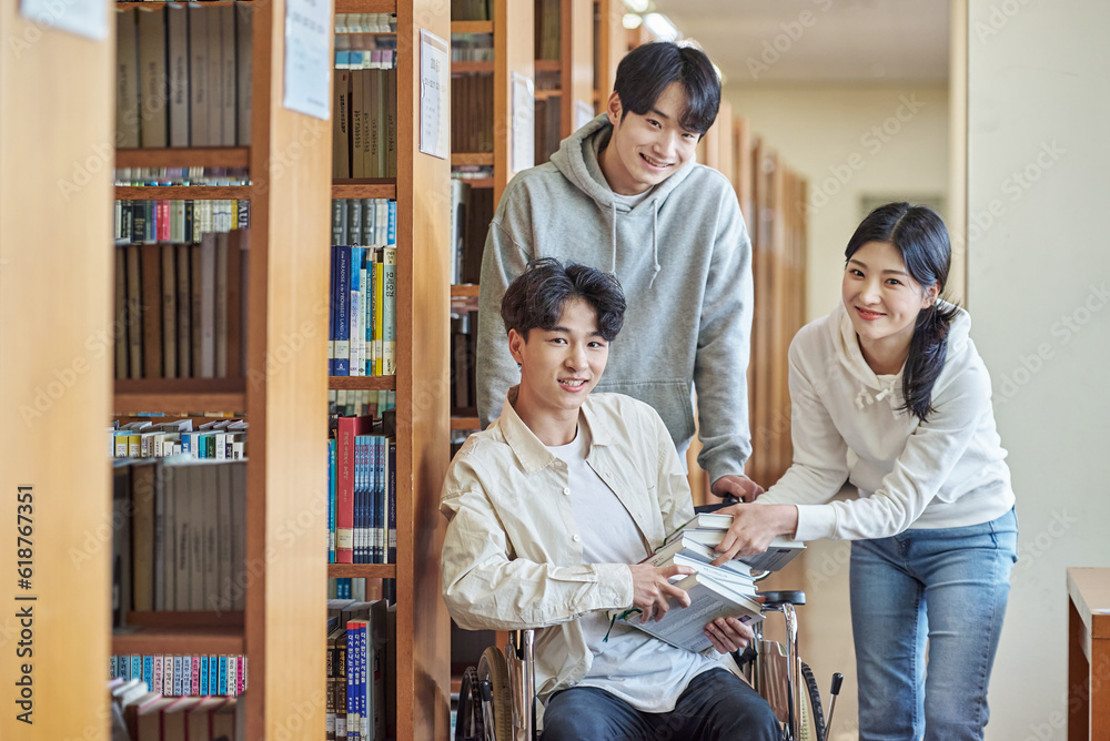 Obraz premium A young male model with a disability sitting in a wheelchair reading a book in a university library in Asia, with two young male and female college students helping him