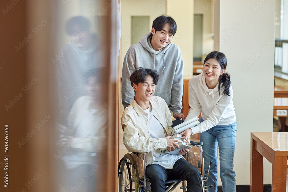 A young male model with a disability sitting in a wheelchair reading a book in a university library in Asia, with two young male and female college students helping him A young male model with a disability sitting in a wheelchair reading a book in a university library in Asia, with two young male and female college students helping him