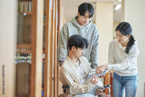 A young male model with a disability sitting in a wheelchair reading a book in a university library in Asia, with two young male and female college students helping him