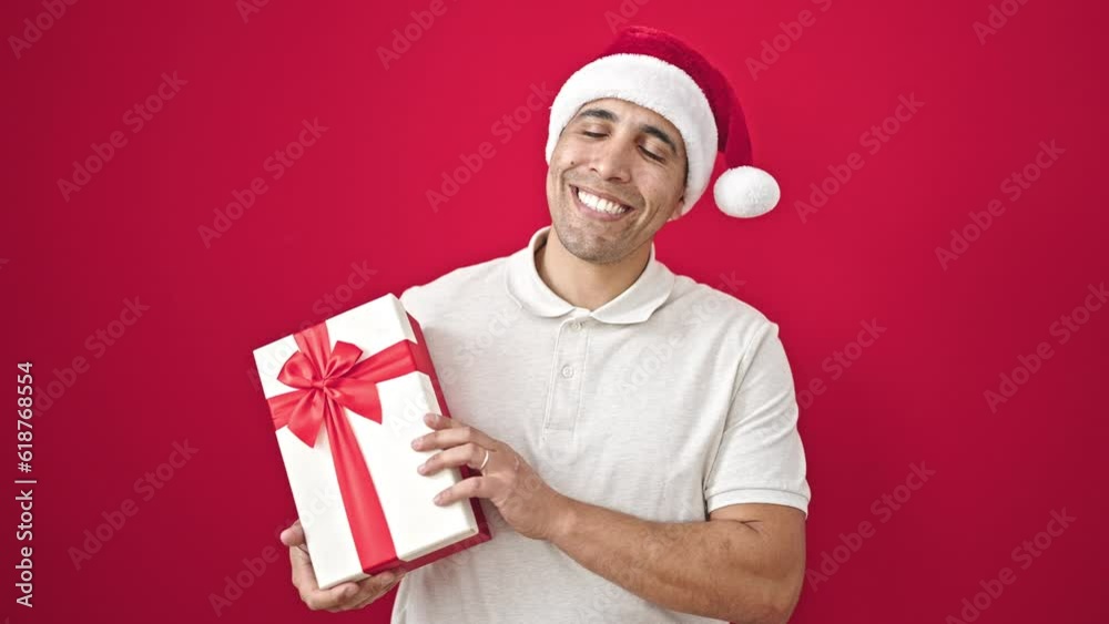 Young hispanic man smiling confident holding christmas present over isolated red background Young hispanic man smiling confident holding christmas present over isolated red background