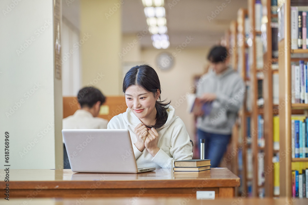 Young Asian Korean female model in library looking at laptop or book, lecture, assignment, discussion, male model in background, bookshelf in background Young Asian Korean female model in library looking at laptop or book, lecture, assignment, discussion, male model in background, bookshelf in background