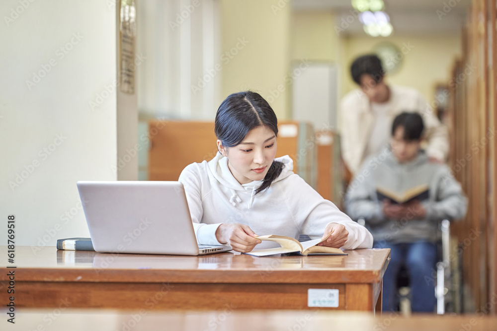 Asian Korean university library desk chair woman sitting on laptop and reading book and man helping disabled man in wheelchair Young Asian male and female models Asian Korean university library desk chair woman sitting on laptop and reading book and man helping disabled man in wheelchair Young Asian male and female models