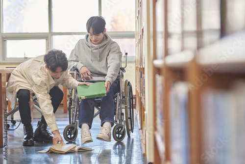 Young handicapped disabled male college student model in wheelchair looking for books in library bookshelf at university in Asia, helping male college student