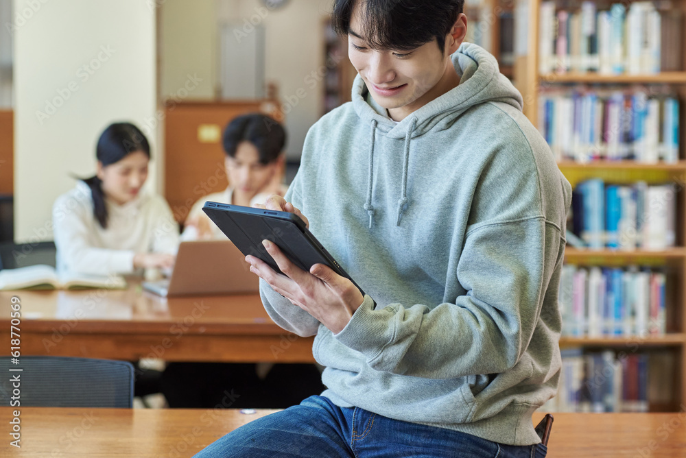 Young Asian Korean male model, man and woman model and looking out the window with tablet in the library on the background of bookshelf Young Asian Korean male model, man and woman model and looking out the window with tablet in the library on the background of bookshelf
