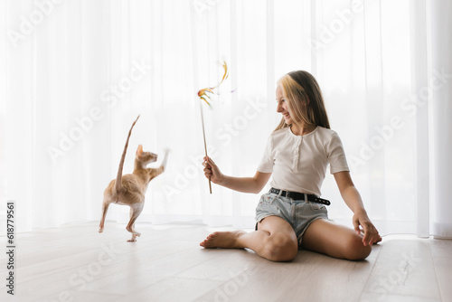 Teen girl sitting on the floor playing with a cat cornish rex with a stick teaser