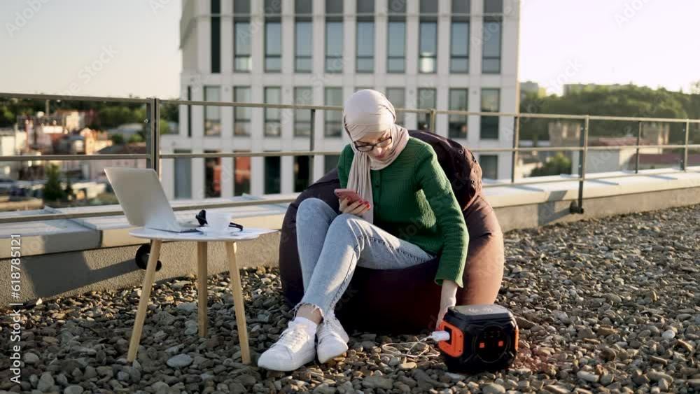 Full length of muslim woman plugging mobile charger into wall outlet of portable power station on panoramic terrace. Relaxed lady in headscarf activating digital device for making business calls. Full length of muslim woman plugging mobile charger into wall outlet of portable power station on panoramic terrace. Relaxed lady in headscarf activating digital device for making business calls.