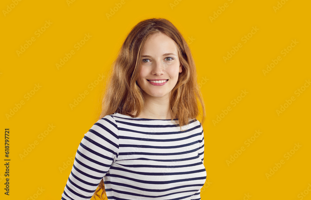 Studio portrait of pretty woman. Headshot of happy beautiful young blonde long haired girl in casual striped top standing isolated on orange yellow background, looking at camera and smiling Studio portrait of pretty woman. Headshot of happy beautiful young blonde long haired girl in casual striped top standing isolated on orange yellow background, looking at camera and smiling