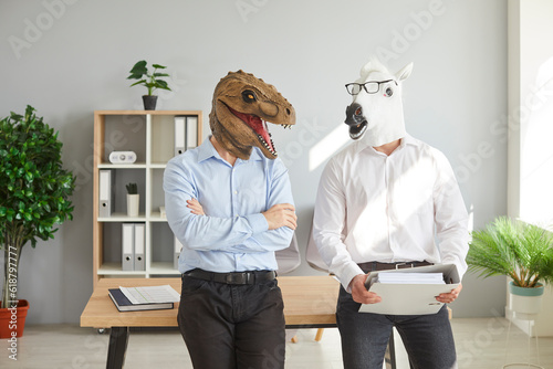 Business people or office workers in funny bizarre animal masks. Two men wearing shirts and unusual absurd dinosaur and horse masks standing by office desk and discussing something. Humor concept