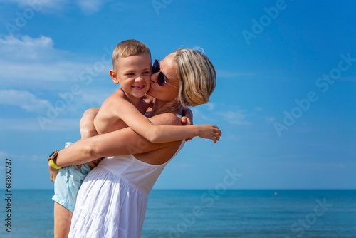 A young mother with a little son hug and kiss on the background of the sea. A woman in a white sundress and sunglasses laughs and plays with her son on the beach. Love and tenderness.