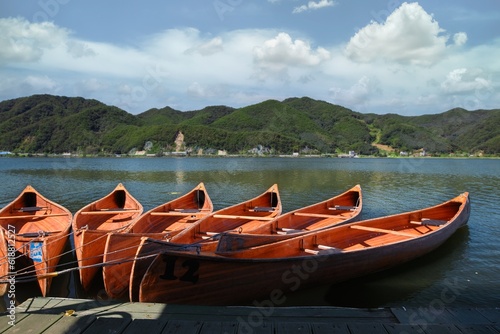 Canoes sit in the water of a river in summer in panorama view. Canoes rest on a bank of a lake, mountains in background. © Emanuele