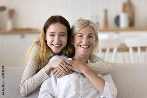 Happy pretty teenage grandkid girl and positive blonde grandmother head shot home portrait. Teen child hugging mature mom from behind, looking at camera with toothy smile, laughing