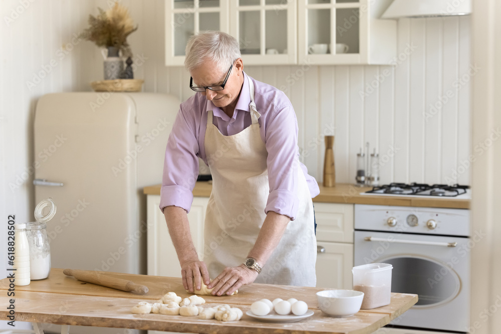 Senior baker man wearing domestic apron patting raw dough on floury table, preparing fresh buns, enjoying culinary hobby, cooking pastry dessert in home kitchen Senior baker man wearing domestic apron patting raw dough on floury table, preparing fresh buns, enjoying culinary hobby, cooking pastry dessert in home kitchen