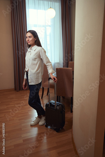 Young lady in hotel holding suitcase in front of window