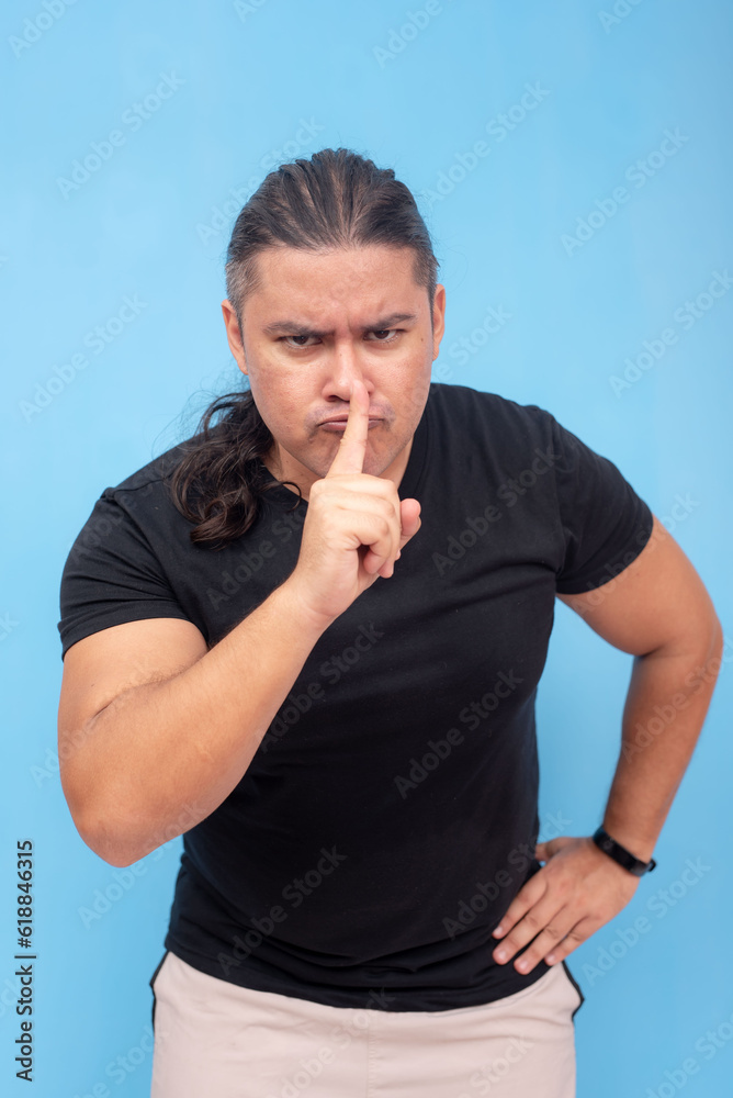 An irate biracial male leaning forward and doing a shush gesture with one finger. Isolated on a blue background. An irate biracial male leaning forward and doing a shush gesture with one finger. Isolated on a blue background.