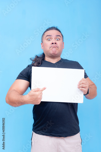 A biracial male with undercut and pursed lips angrily holding a blank placard while pointing at it with one hand. Isolated on a blue background.