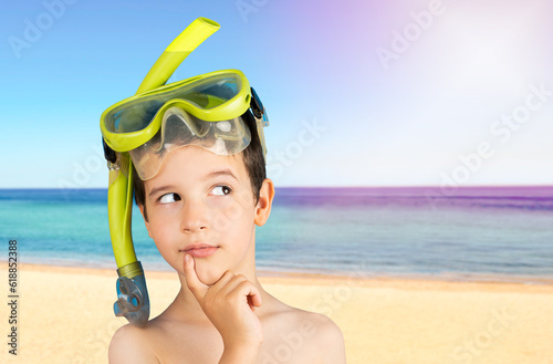 Pensive child with snorkel mask tuba and snorkel looks away at copyspace thinking at beach,funny kid lips hold finger near mouth conceiving some kind of joke.Snorkeling, swimming, vacation concept