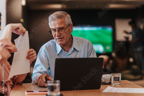 70s experienced male business owner looking at lap top reading achievements, work flow and efficiency and profit growth, discussing at cafe at modern office