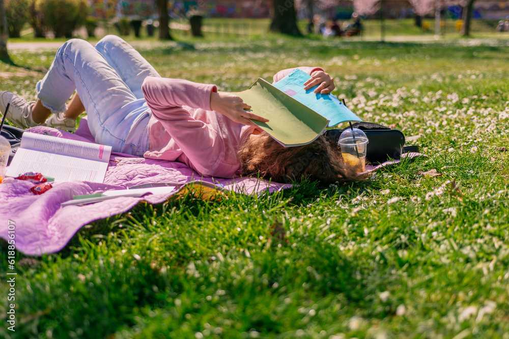 Blonde curly haired girl lying on blanket at the park with book covering her face, protecting from the sun