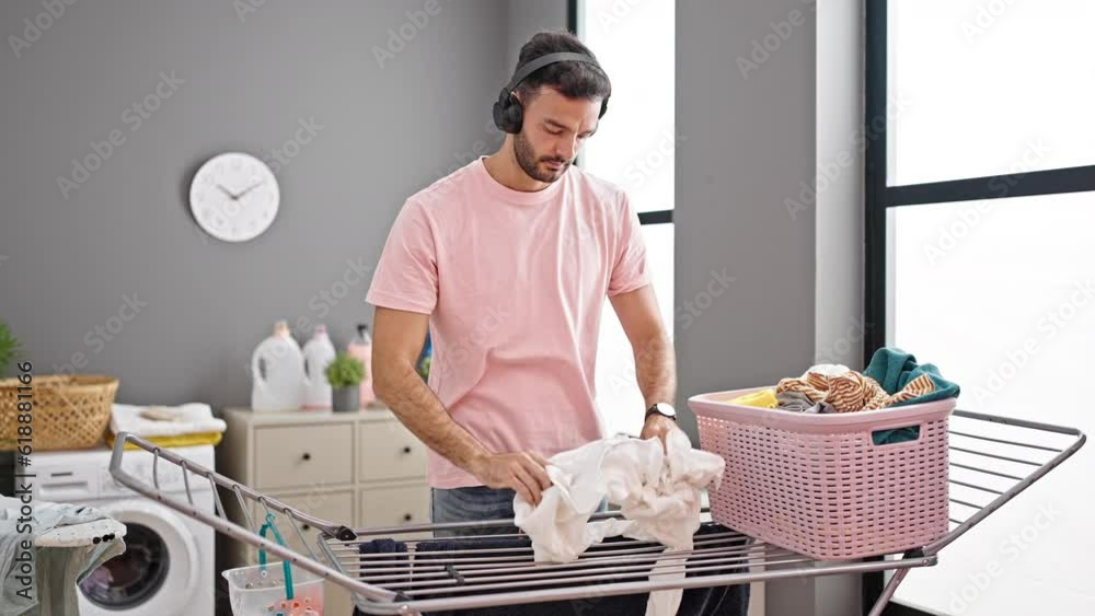 Young hispanic man listening to music hanging clothes on clothesline at laundry room