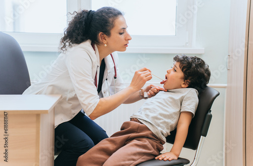 Serious brunette Spanish female doctor checks throat of little  boy at medical office. Pandemic concept. Toddler at medical exam at hospital. Healthcare, healthy  childhood. photo