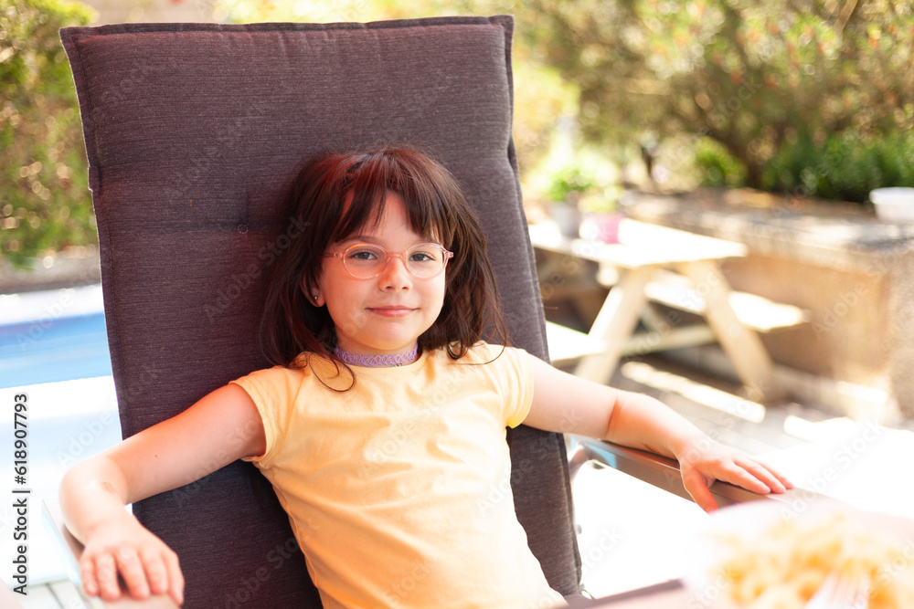 Naklejka premium Cheerful little laughing girl in glasses sits in a chair in a cafe on a warm summer day