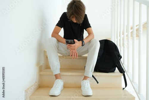 Teenage boy using smartphone. Unhappy, sad, frustrated teenage boy sitting alone on stairs. Learning difficulties, gaming addiction, emotion, psychological problems in adolescence concept