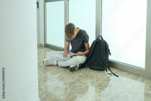 Teenage boy student wearing casual clothes sitting on floor, looking to smartphone and writing, makes notes in notebook. Teenager studies, learning, writing, doing homework, preparing to exam