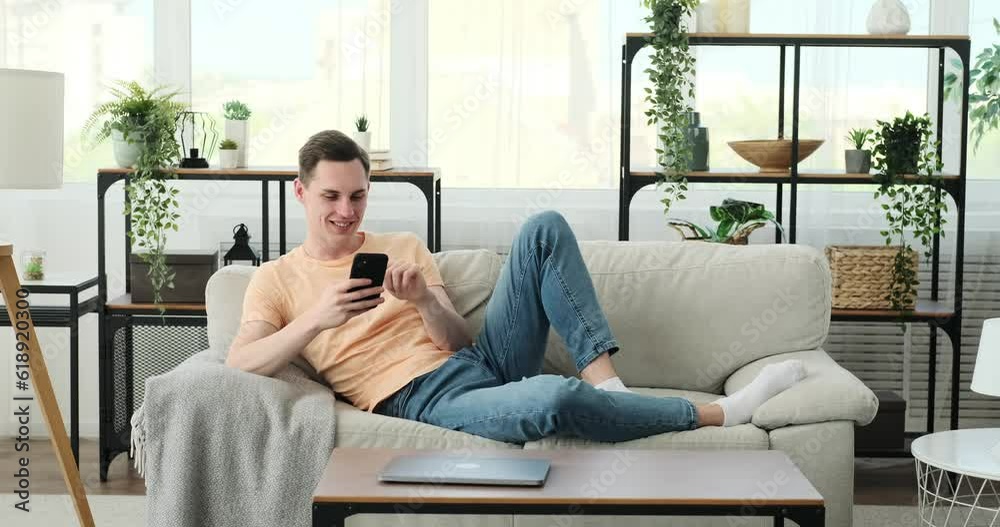 Joyful Caucasian man as he engages in lively conversations and bursts into laughter while using his phone on the sofa. With a bright smile on his face and an unmistakable twinkle in his eyes. Joyful Caucasian man as he engages in lively conversations and bursts into laughter while using his phone on the sofa. With a bright smile on his face and an unmistakable twinkle in his eyes.