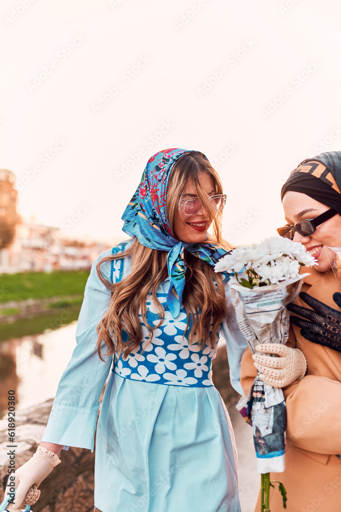 Couple woman one wearing a hijab and a modern yet traditional dress, and the other in a blue dress and scarf, walking together through the city at sunset. One carries a bouquet and bread, while the