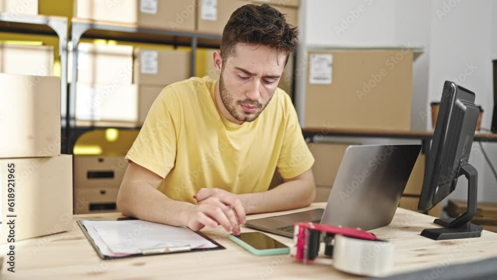 Young hispanic man ecommerce business worker using smartphone and laptop writing on document at office