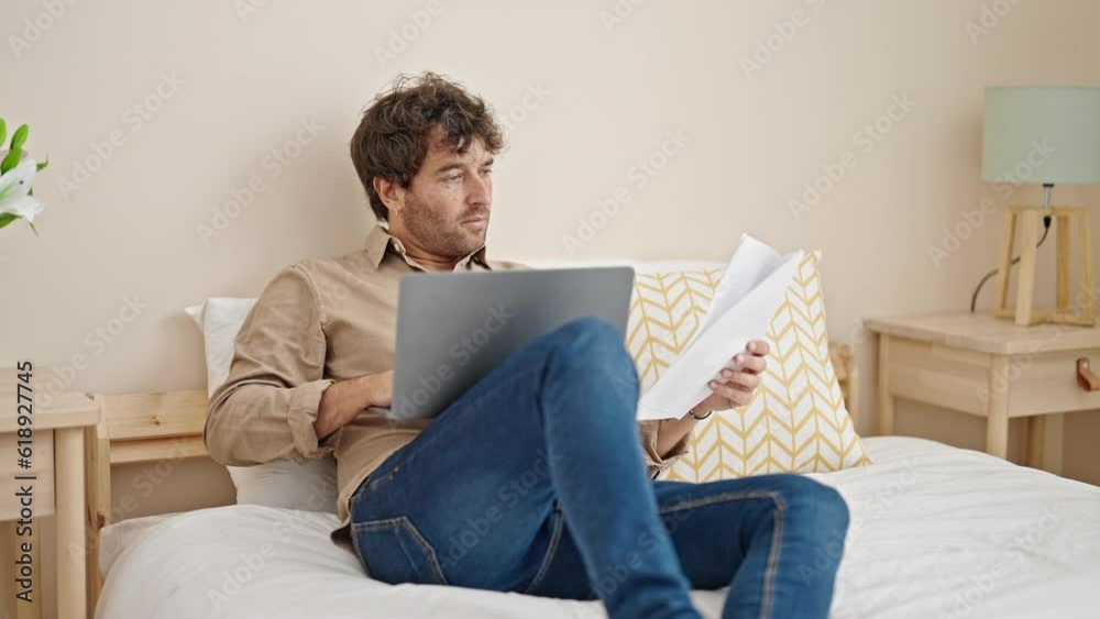 Young hispanic man using laptop reading document sitting on bed at bedroom Young hispanic man using laptop reading document sitting on bed at bedroom