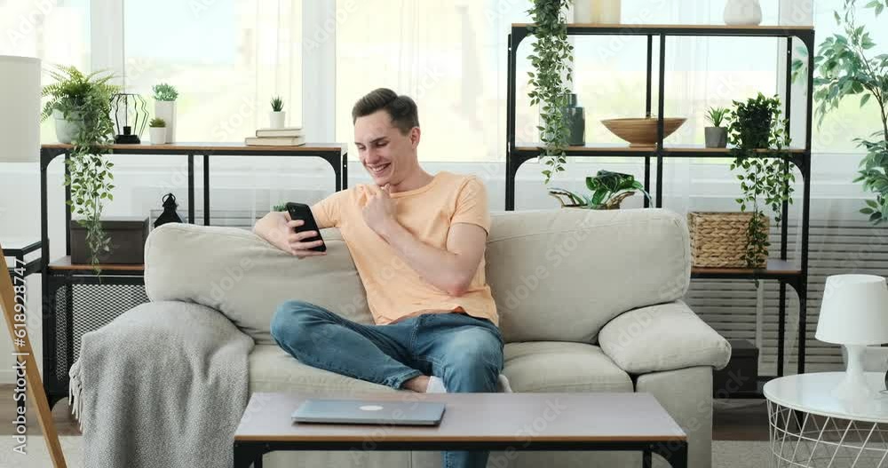 Man seated on the couch, his face adorned with a genuine smile, and his attention captivated by the phone in his hands. Completely at ease, he indulges in the digital realm, letting go of any worries. Man seated on the couch, his face adorned with a genuine smile, and his attention captivated by the phone in his hands. Completely at ease, he indulges in the digital realm, letting go of any worries.