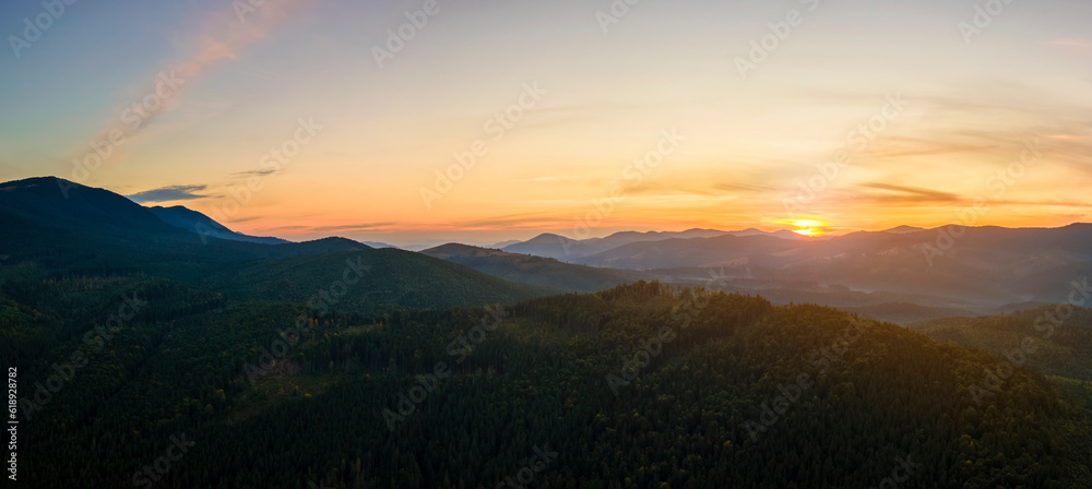 Obraz premium Aerial view of dark mountain hills with bright sunrays of setting sun at sunset. Hazy peaks and misty valleys in evening