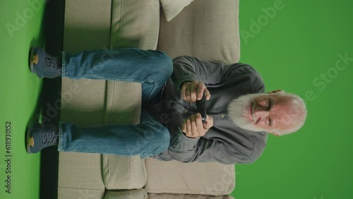 Vertical View.Green Screen. An Old Man With a Gray Beard Plays Computer Games With a Joystick. An Elderly Man Plays PlayStation and Wins.Tech Nostalgia and Generational Divide. photo