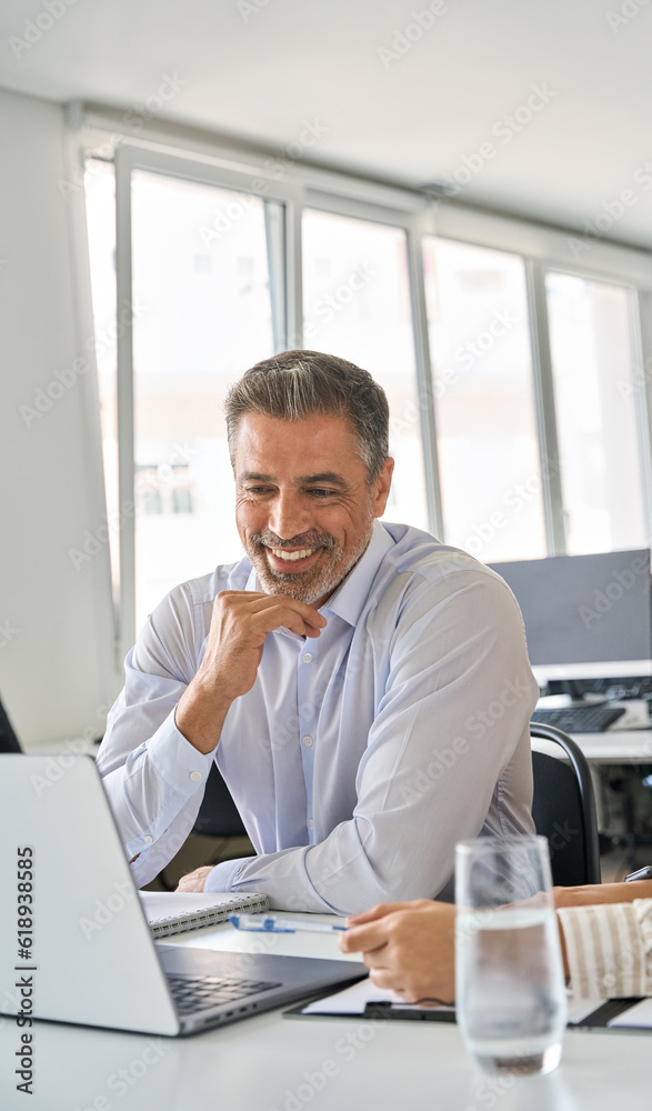 Fototapeta premium Happy professional mid aged mature Latin executive manager working in office with female colleague, talking and looking at laptop computer sitting at table workplace. Vertical authentic shot.