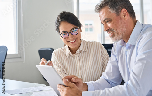 Two happy professional business people team Asian woman and Latin man workers working using digital tablet tech having conversation discussing financial marketing strategy at corporate office meeting.