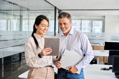 Canvas Print Two happy professional business people team Asian woman and Latin man workers working using digital tablet tech discussing financial market data standing at corporate office meeting
