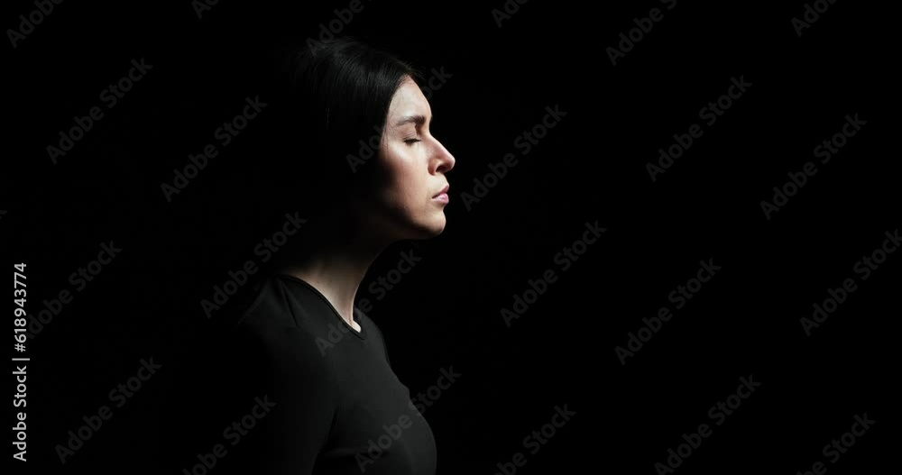 Woman standing in profile on a black background, breathing deeply and dramatically lifting her head to gaze upwards. With a serious expression and a thoughtful demeanor.