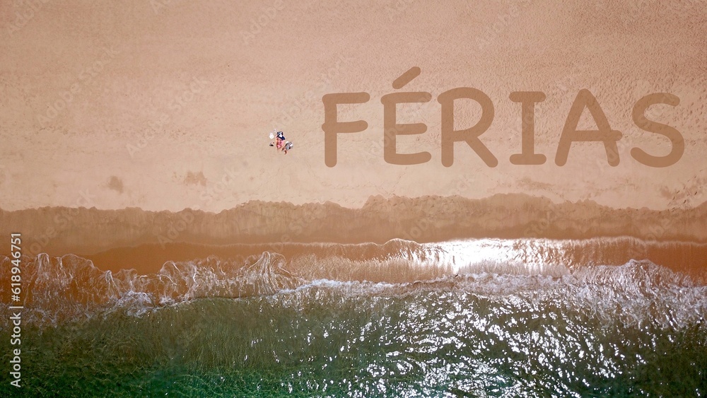 Vista aérea de uma bela praia de areia junto ao mar azul, onde dois veranistas estão deitados e tomando banho de sol, ao lado deles a inscrição "FÉRIAS" na areia - obrazy, fototapety, plakaty 