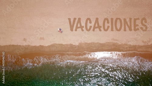 Vista aérea de una hermosa playa de arena junto al mar azul, donde dos vacacionistas yacen y toman el sol, junto a ellos las letras 