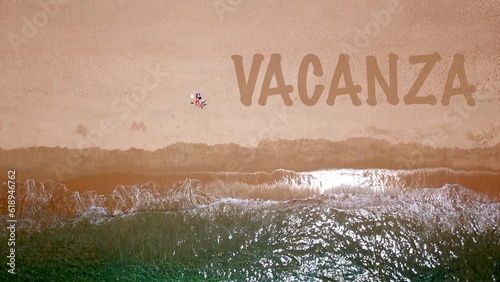 Vista aerea di una bellissima spiaggia sabbiosa in riva al mare blu, dove due vacanzieri sono sdraiati e prendono il sole, accanto a loro la scritta 