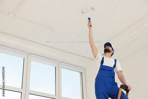 Worker in uniform painting ceiling with roller indoors, low angle view