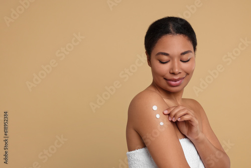Young woman applying body cream onto shoulder on beige background. Space for text