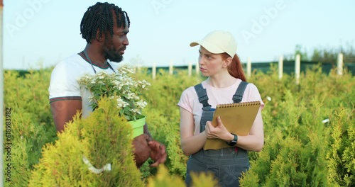 African American garderner giving advise to Caucasuan redhead woman how to grow plants, take care of bushes, flowers in greenhouse, man sharing secrets of successful cultivation garderning Slow motion photo