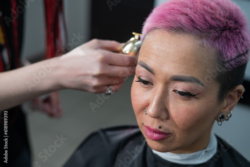 The hairdresser shaves the temple of a female client. Asian woman with short pink hair in barbershop.